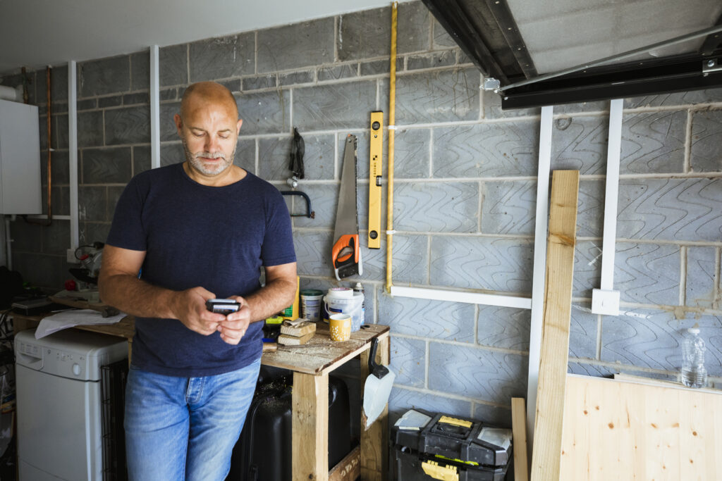 A man is using his mobile phone to surf the web while standing in his garage. He is leaning on the workbench beside him. Work tools and cut wood can be seen on the bench.